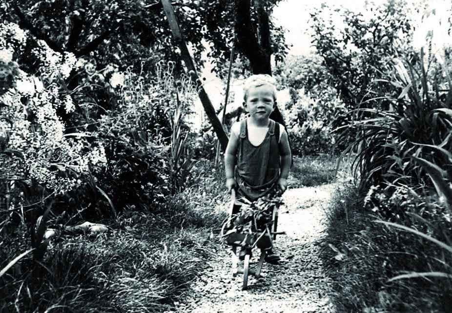 A smiling child pushes a tricycle in a lush garden, surrounded by flowers and foliage. A smiling child pushes a tricycle in a lush garden, surrounded by flowers and foliage.