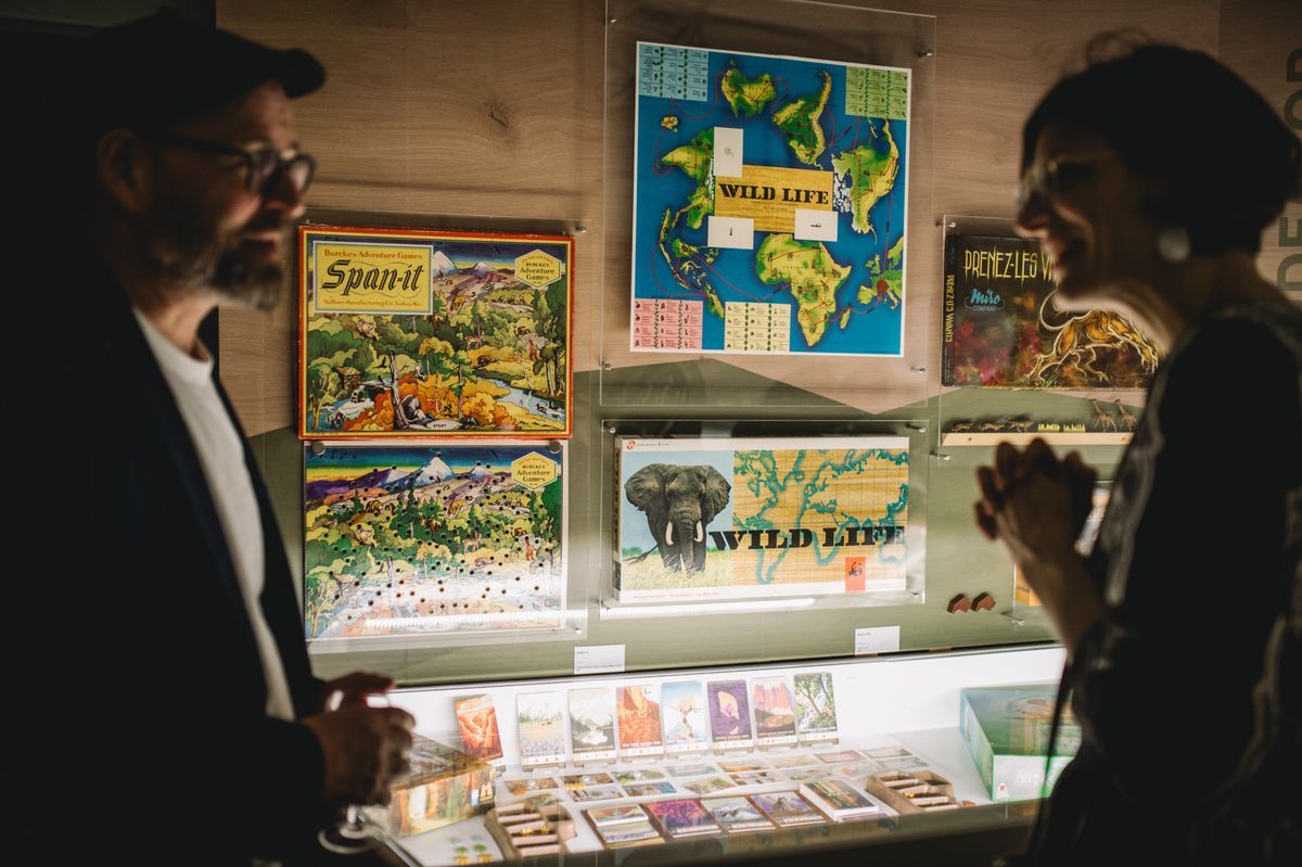 Two people chat in front of a showcase containing board games and educational cards, lit by hot light. Two people chat in front of a showcase containing board games and educational cards, lit by hot light.