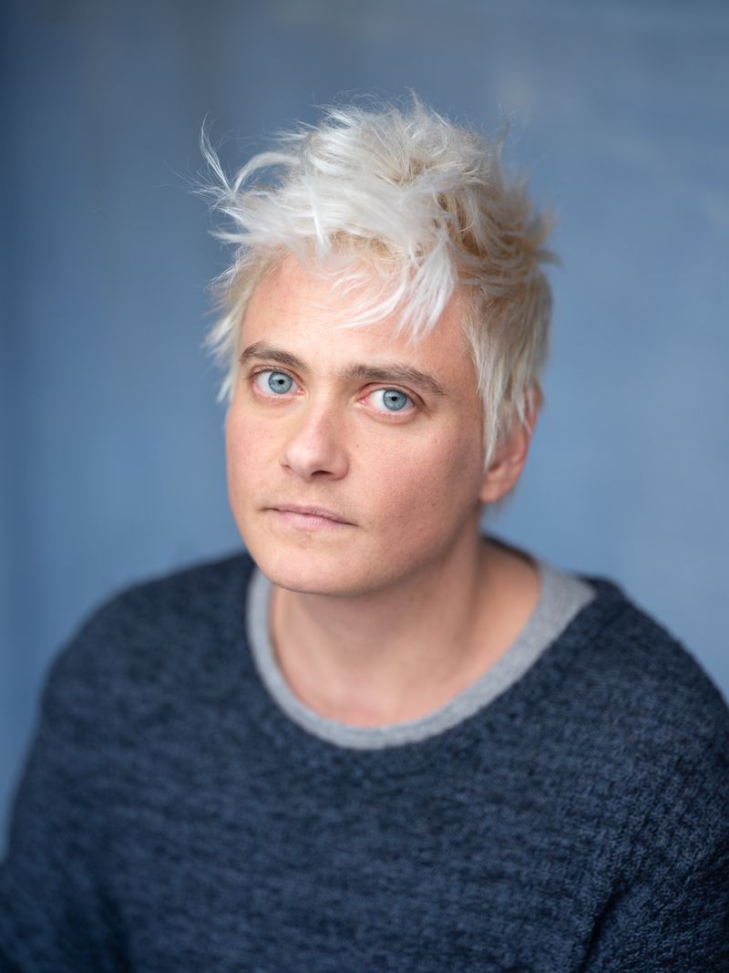 Person with light hair wearing a gray sweater and looking forward with a light blue background. Person with light hair wearing a gray sweater and looking forward with a light blue background.