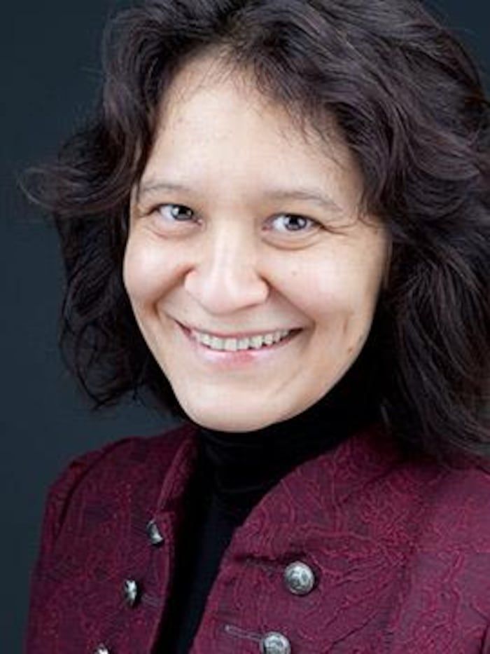 A woman with brown hair, smiling, wearing a red jacket and a black turtleneck, pose.