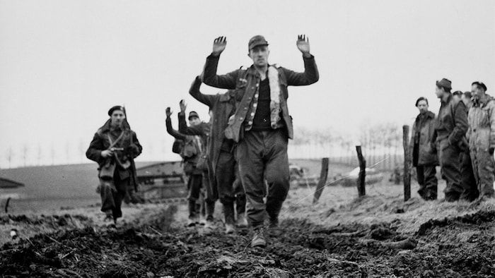 Photo of archives showing a soldier, a hand weapon guiding prisoners, hands in the air.