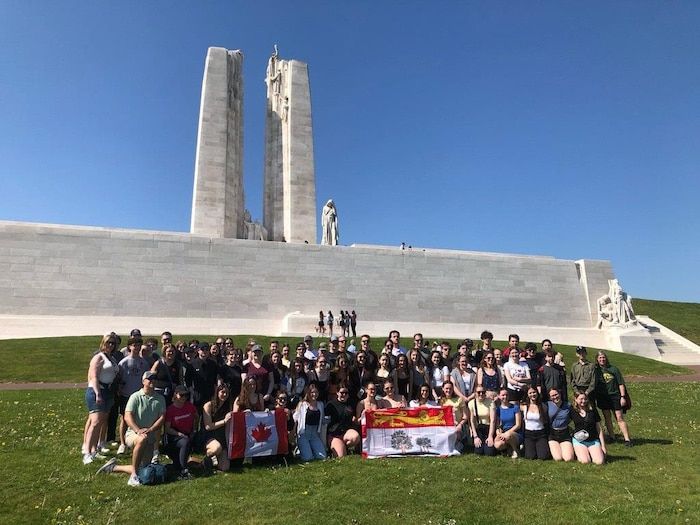 Dozens of adolescents are kneeling or standing in the grass with a Canada flag and one of the Prince Edward Island, with the Vimy monument back.