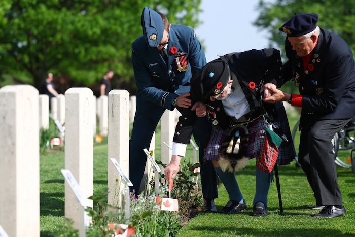 A centenary in military uniform decreases to deposit something at the foot of a grave, while a young soldier and another older man help him.
