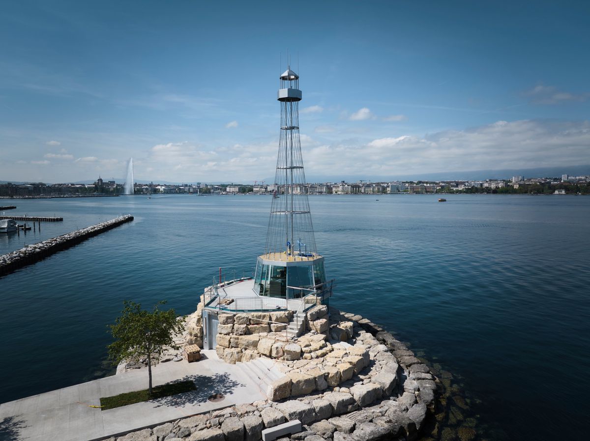 View of a modern lighthouse overlooking Lake Geneva in Geneva by a sunny day, with the city visible in the background. View of a modern lighthouse overlooking Lake Geneva in Geneva by a sunny day, with the city visible in the background.