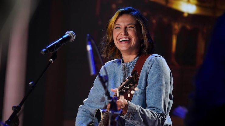 Sara Dufour smiles in front of a microphone with her guitar in hand.
