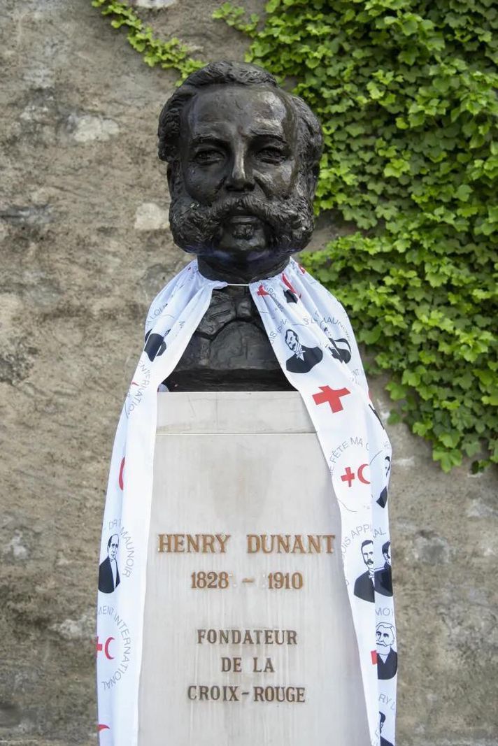 Bust of Henry Dunant covered with an illustrated scarf, founder of the Red Cross, with a stone wall and greenery in the background. Bust of Henry Dunant covered with an illustrated scarf, founder of the Red Cross, with a stone wall and greenery in the background.