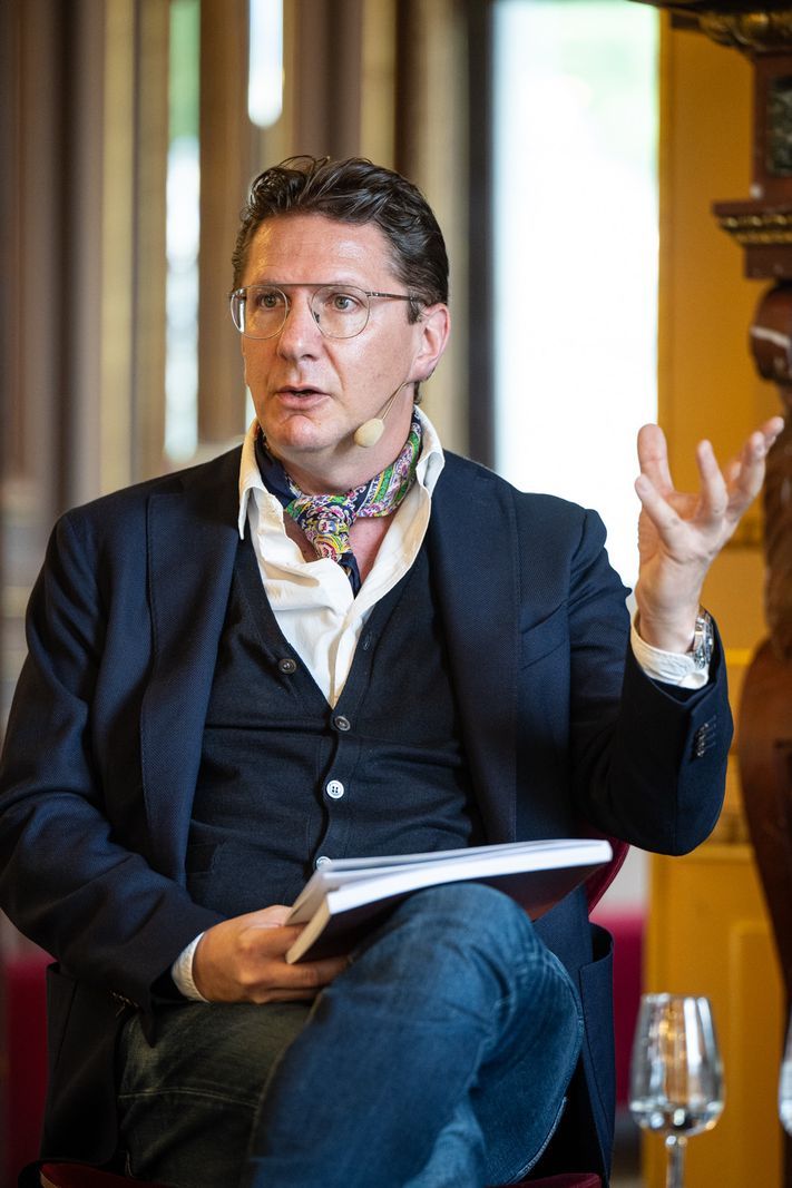Man in costume with scarf, holding a notebook and speaking during an event. Man in costume with scarf, holding a notebook and speaking during an event.