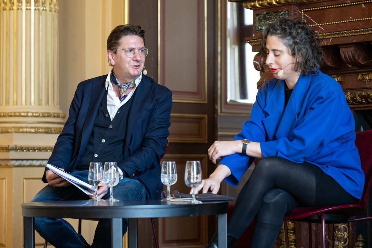 Two people chatting sitting at a round table, with glasses of water in front of them, in an elegant room with golden columns. Two people chatting sitting at a round table, with glasses of water in front of them, in an elegant room with golden columns.