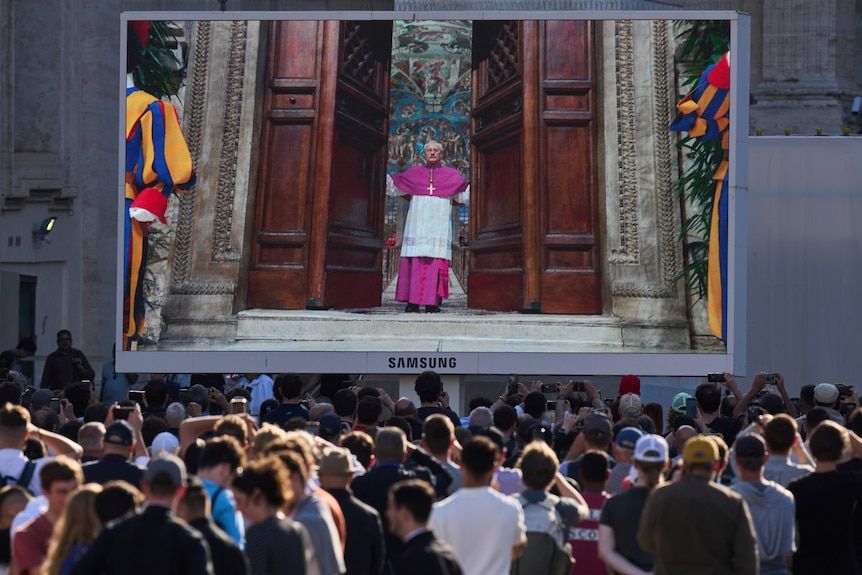 A giant Samsung TV screen showing a Cardinal in purple and white robes shutting large brown wooden doors