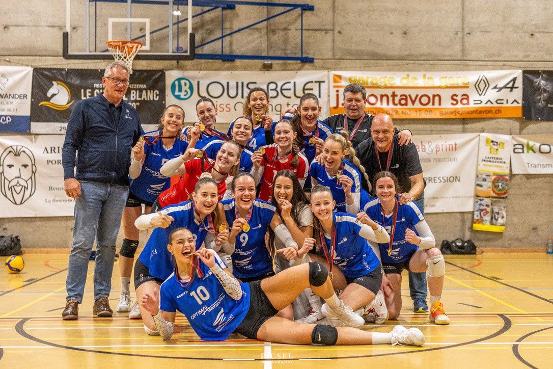 Volleyball women's team celebrating a victory, smiling with medals around the neck, surrounded by advertising banners in a gymnasium. Volleyball women's team celebrating a victory, smiling with medals around the neck, surrounded by advertising banners in a gymnasium.
