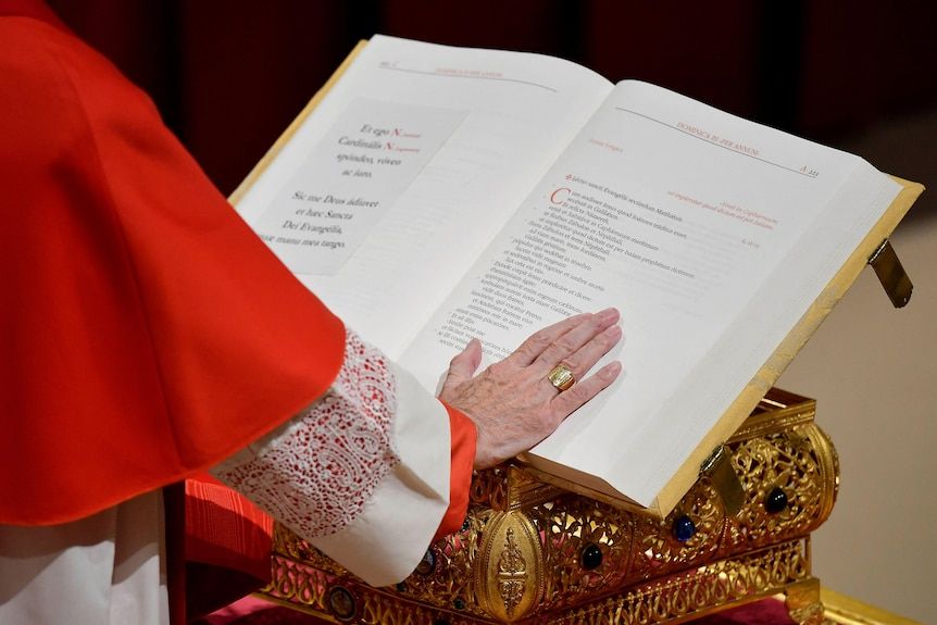 A Cardinal's sleeve in a red and white gown with one palm on an open prayer book on a golden lectern