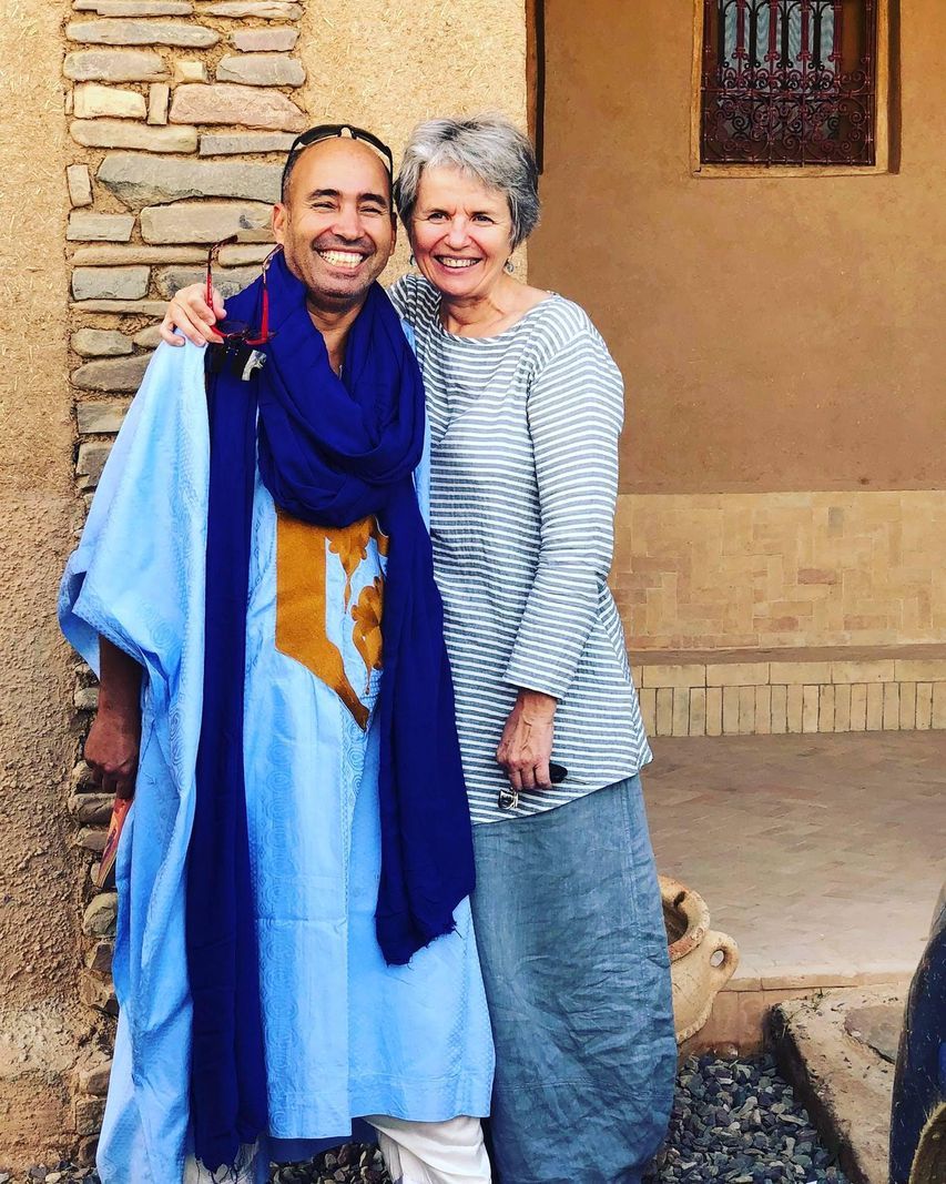 Two smiling people posing together in front of a stone building, one wearing a traditional blue garment. Two smiling people posing together in front of a stone building, one wearing a traditional blue garment.