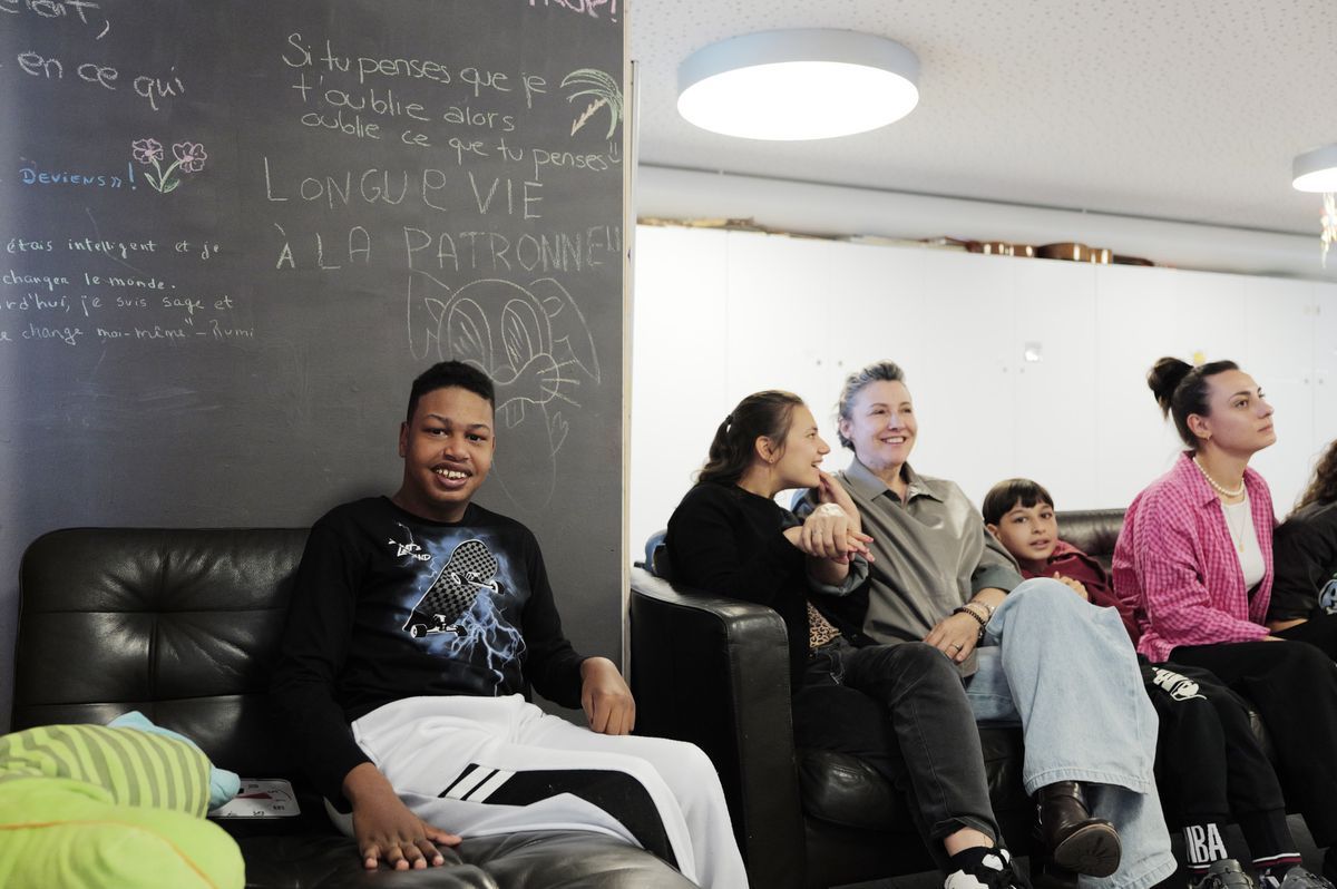 Young people sitting in a living room at the Clair Bois Foundation in Geneva, chatting and smiling, with a blackboard in the background showing drawings and inscriptions. Young people sitting in a living room at the Clair Bois Foundation in Geneva, chatting and smiling, with a blackboard in the background showing drawings and inscriptions.