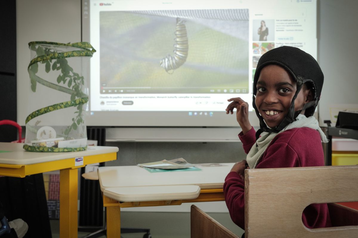 A child sitting at a table in a classroom of the Clair Bois Foundation in Plan-les-Ouates, smiling on the camera with a protective helmet. In the background, a screen projects a video on the caterpillars, in connection with the open door event on childhood and adolescence. A child sitting at a table in a classroom of the Clair Bois Foundation in Plan-les-Ouates, smiling on the camera with a protective helmet. In the background, a screen projects a video on the caterpillars, in connection with the open door event on childhood and adolescence.